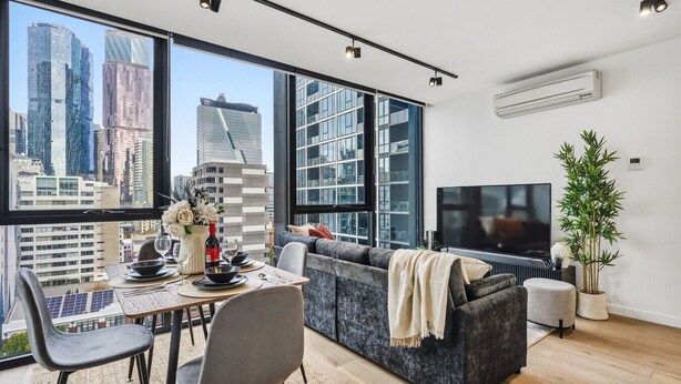 A modern living room with a dining area, large windows, a flat-screen TV, and a potted plant. - CBD-Edge 2BR | Pool, Gym, Sauna & City Views (West Melbourne)
