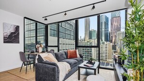 A modern living room with a sofa, coffee table, and dining area, large windows with a city view, and a potted plant. - CBD-Edge 2BR | Pool, Gym, Sauna & City Views (West Melbourne)