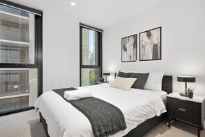 A modern bedroom with a large bed, two bedside tables, a window, and abstract wall art. - CBD-Edge 2BR | Pool, Gym, Sauna & City Views (West Melbourne)