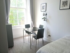 Une pièce comprenant un lit, une table avec des chaises, une fenêtre avec des rideaux, une plante fixée au mur et un tableau encadré.