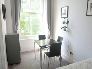 Une pièce avec une fenêtre, un bureau, deux chaises, un lit, une petite armoire et une lampe fixée au mur.