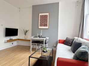Ein modernes Wohnzimmer mit Flachbildfernseher, einem Esstisch mit Stühlen, einem Glastisch und einer Couch mit Kissen.