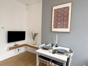 En moderne stue med et fladskærms-tv, et spisebord dækket med tallerkener og glas, en vase med grene, og et blomsterbillede i ramme på væggen.
