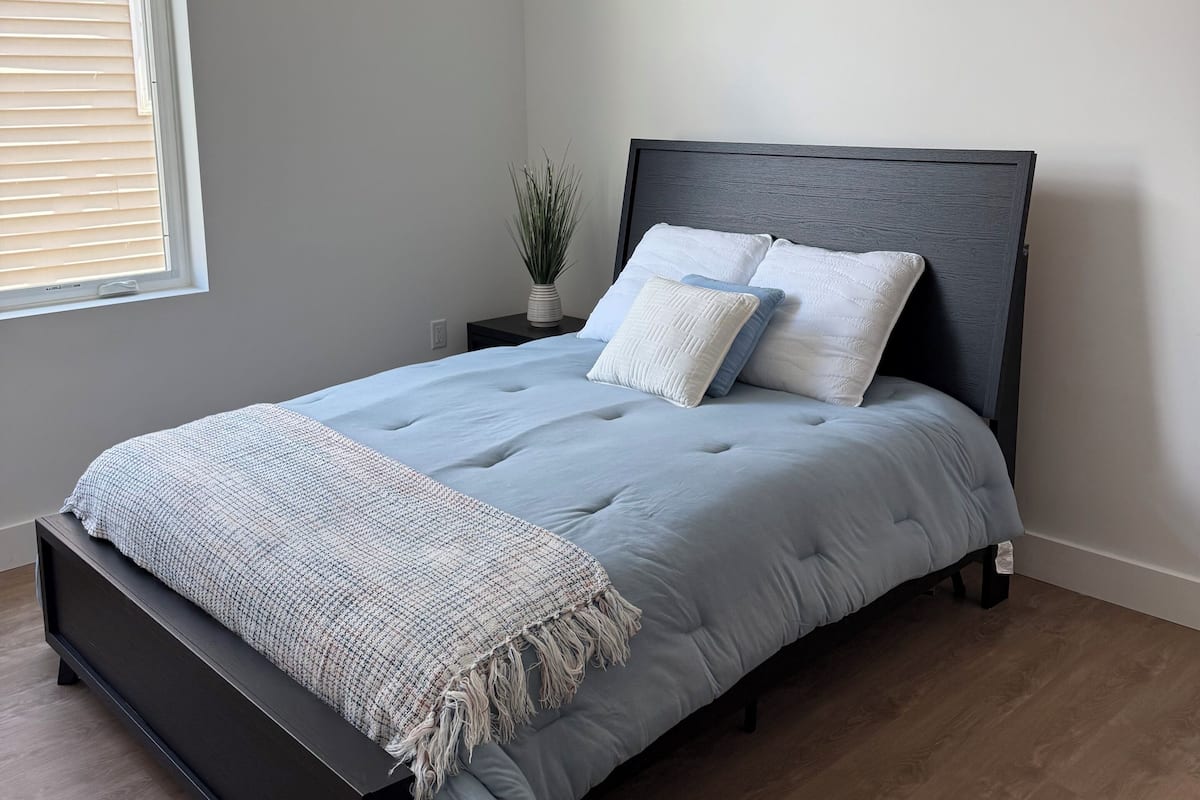 Ein Schlafzimmer mit einem Bett, einem Deckenventilator, einem Fenster mit Jalousien, einer kleinen Pflanze und einem Nachttisch.