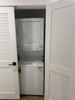 A laundry room with a front-loading washer and dryer, a door with a handle, and a control panel with various buttons and dials. - Coral Sands (Seaside Heights)