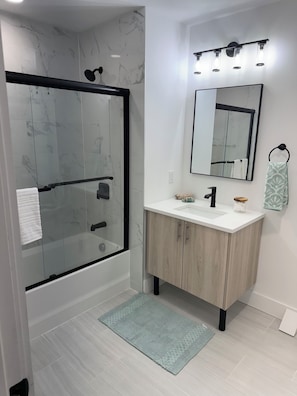 Un baño moderno con ducha envuelta en vidrio, tocador blanco con espejo y alfombra azul claro.