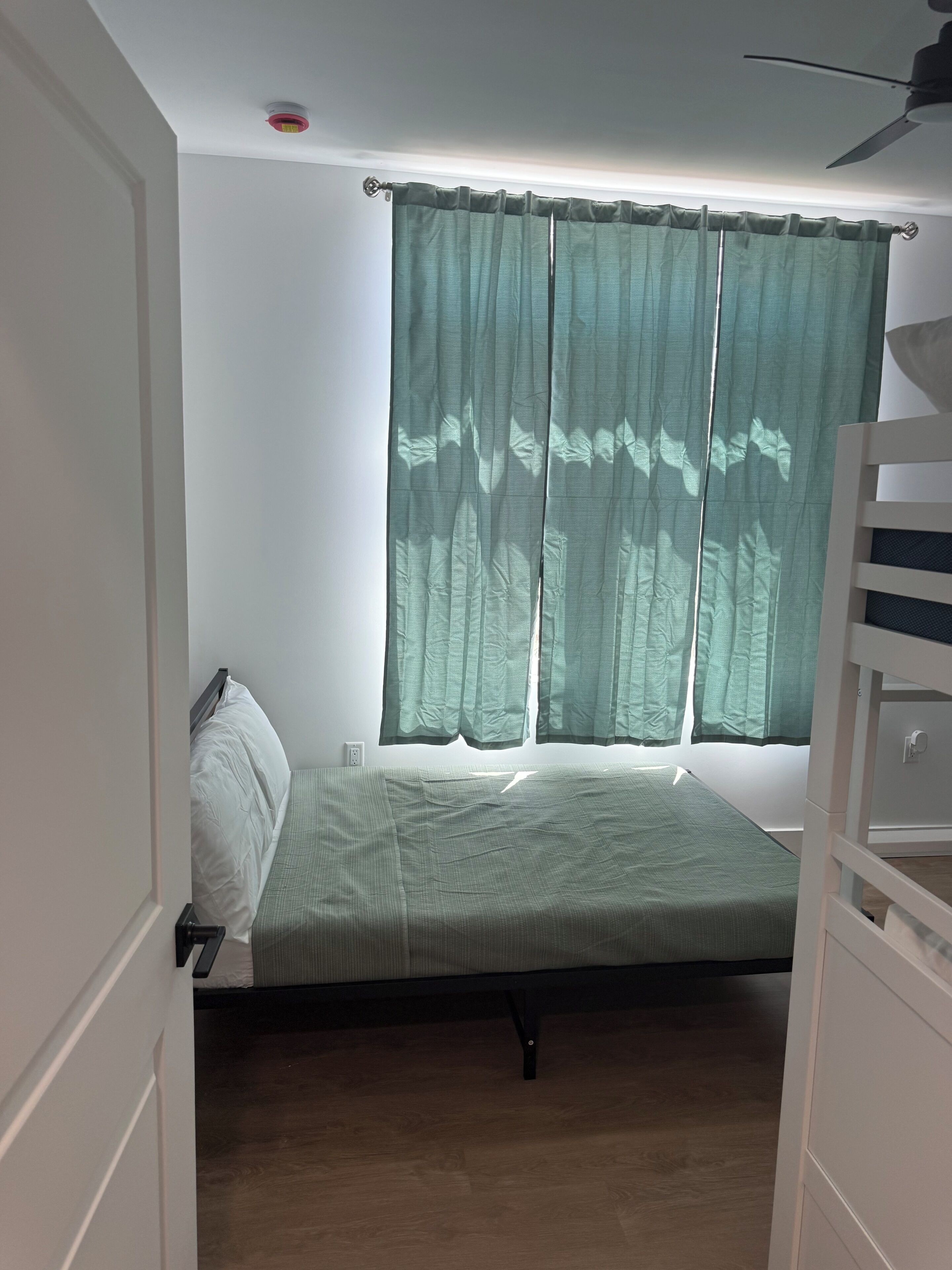 A room with a bed, a window with curtains, a ceiling fan, and a door.