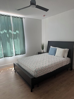 A bedroom with a bed, a ceiling fan, a window with curtains, a nightstand, and a potted plant. - Coral Sands (Seaside Heights)