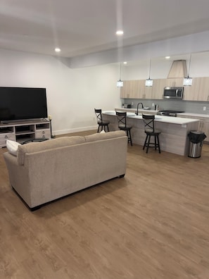 A modern living room with a sofa, a flat-screen TV, and a kitchen area with wooden cabinets and a microwave. - Coral Sands (Seaside Heights)
