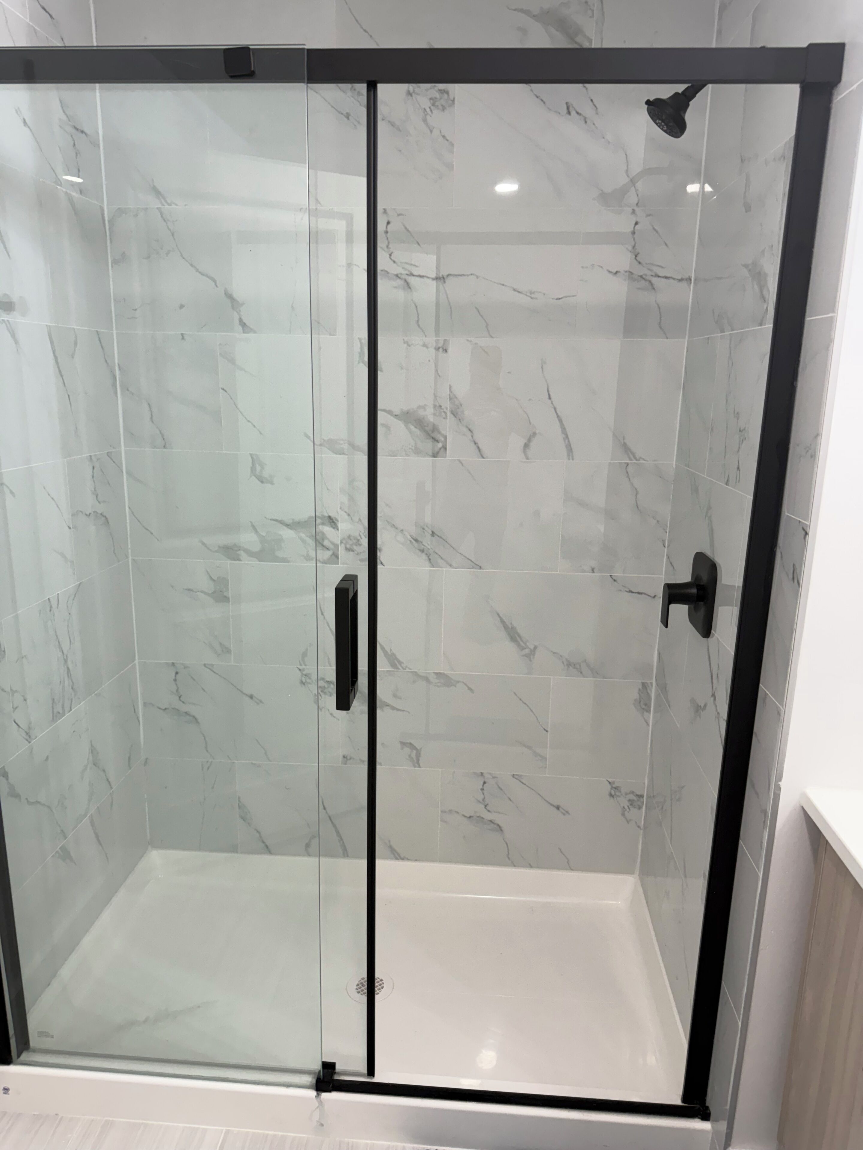 Une cabine de douche fermée par des parois de verre, avec des carreaux de marbre et des accessoires noirs.