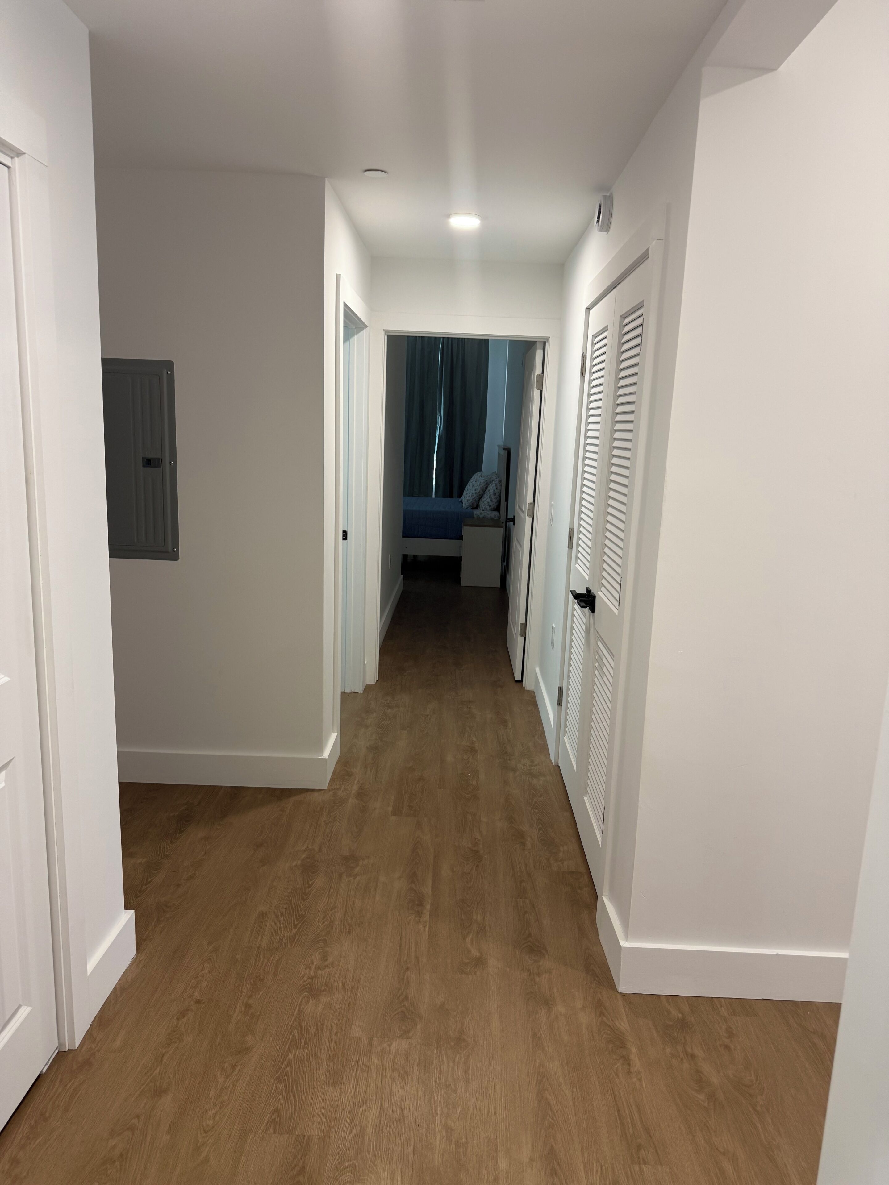 Un couloir doté d’un plancher en bois, de murs blancs et d’une porte donnant sur une chambre à coucher avec un lit et des rideaux.