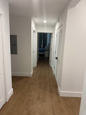 Un pasillo con piso de madera, paredes blancas y una puerta que da a un dormitorio con cama y cortinas.