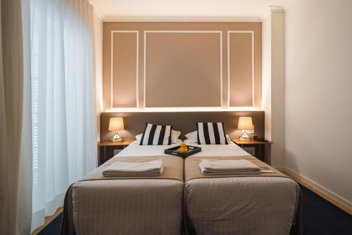 A hotel room with a double bed, two bedside tables with lamps, a tray with a bottle, and a window with curtains.