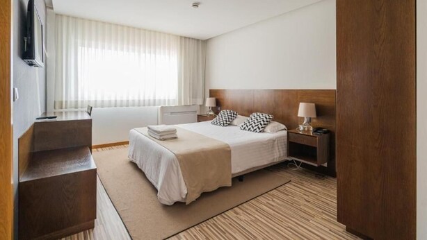A hotel room with a bed, bedside tables, a TV, and a window with curtains. - Residencial Farol (Ilhavo)