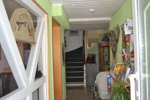 Un pasillo estrecho con una escalera, una puerta con un detalle decorativo y una mesa desordenada con diversos objetos.