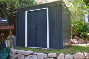 A small, dark-colored shed with a white trim, a stone wall in front, and a garden area with rocks and greenery. - Mahret (Eisenach)