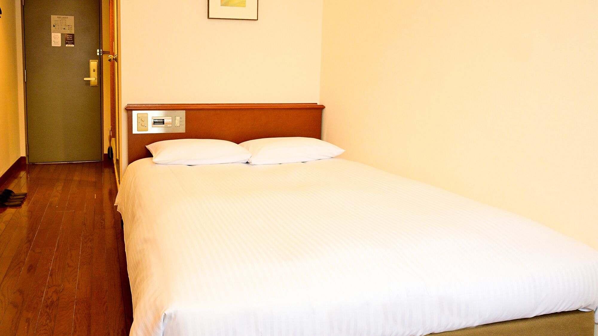 A hotel room with a wooden floor, a bed with white bedding, a wall-mounted control panel, and a door with a sign.