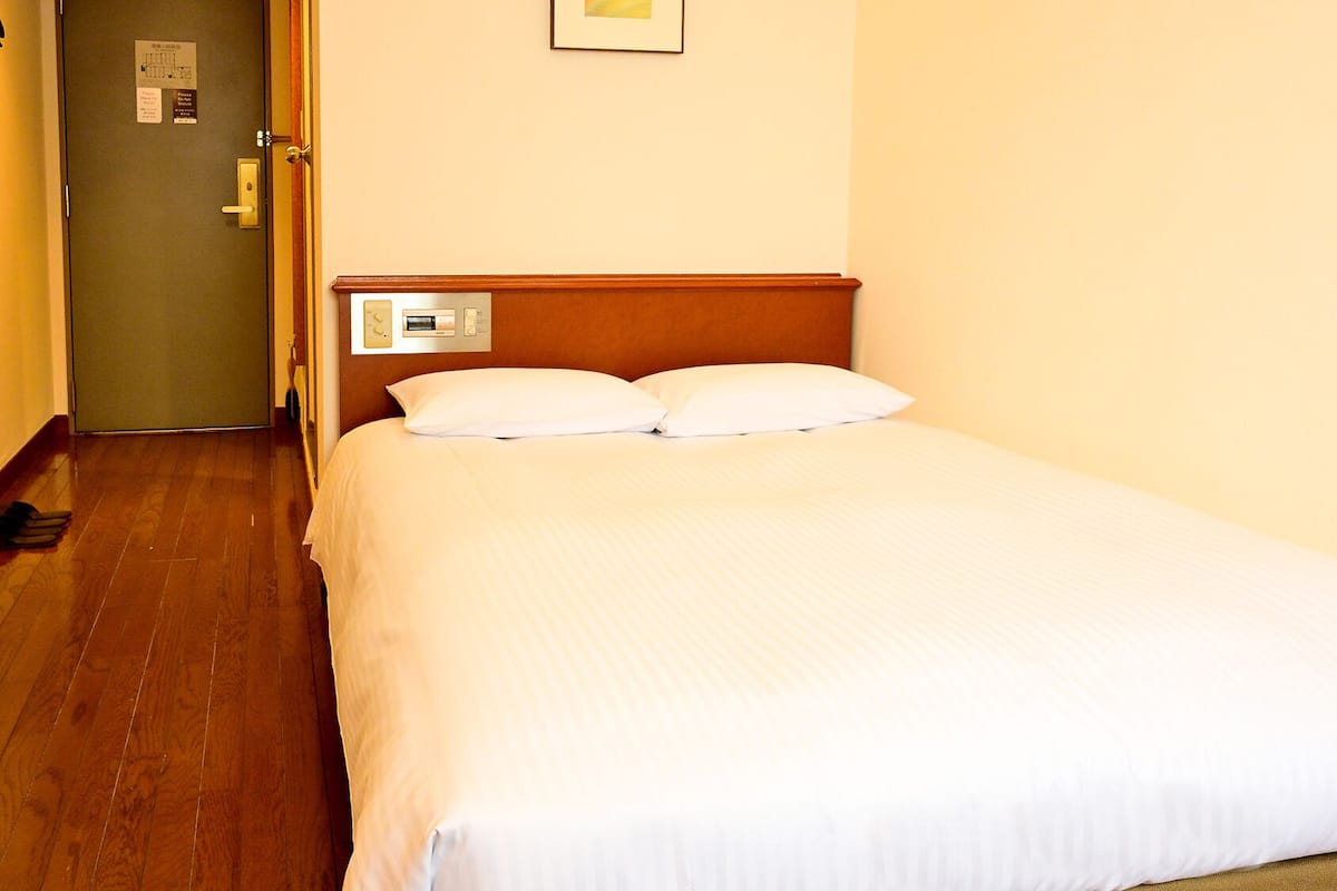 Habitación de hotel con piso de madera, cama con ropa de cama blanca, panel de control en la pared y una puerta con un letrero.