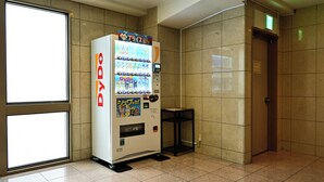 Un distributeur automatique marqué « DyDo » se trouve dans un couloir aux murs carrelés, près d’une porte.