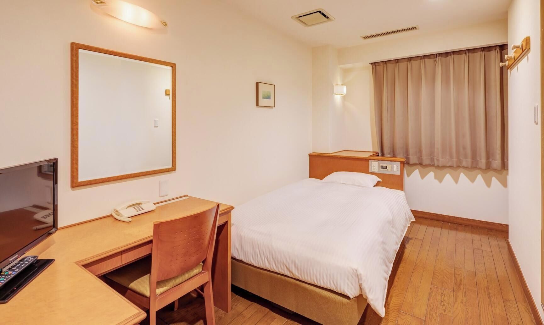 A hotel room with a bed, desk, chair, bedside table, lamp, and a window with curtains.