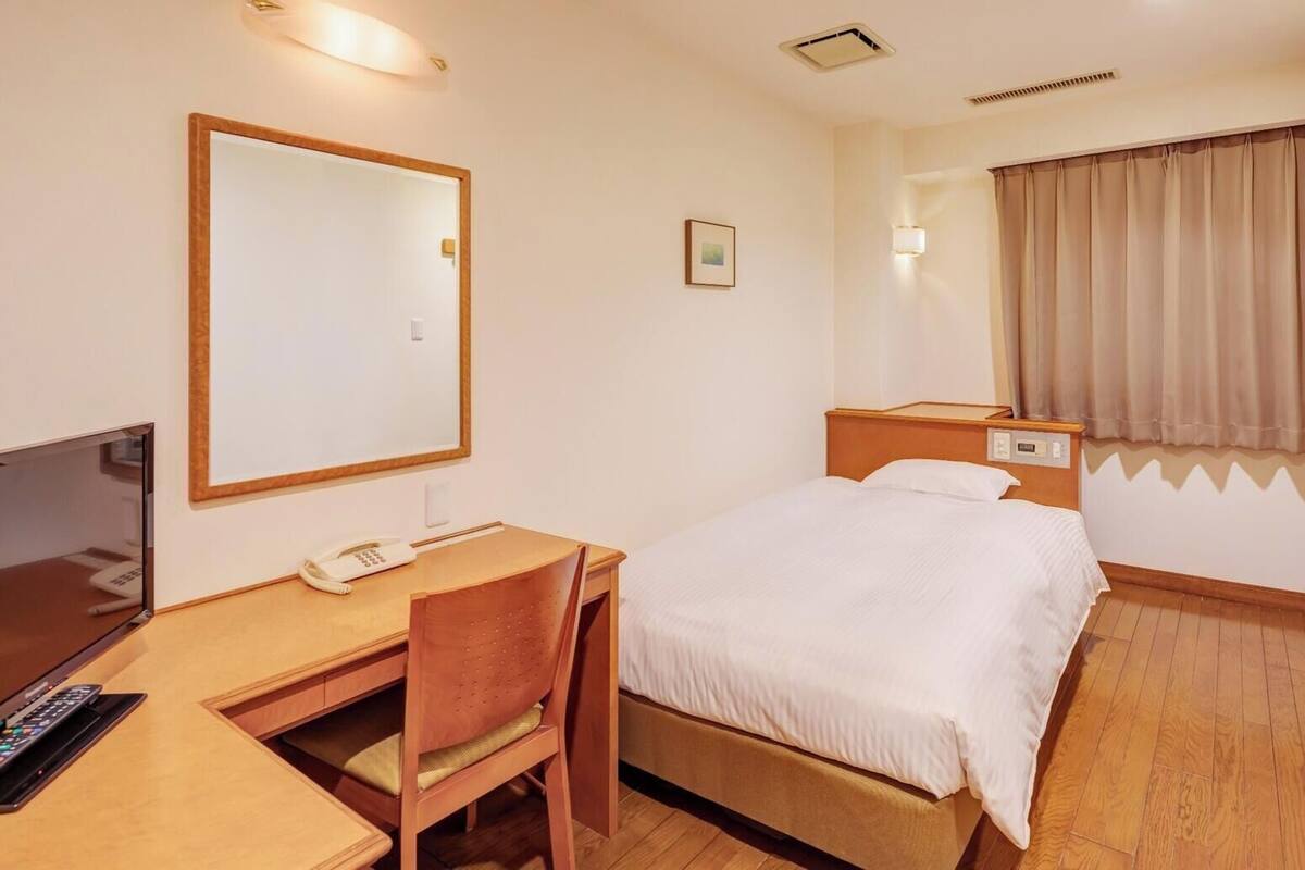Habitación de hotel con cama, escritorio, silla, mesita de noche, lámpara y ventana con cortinas.