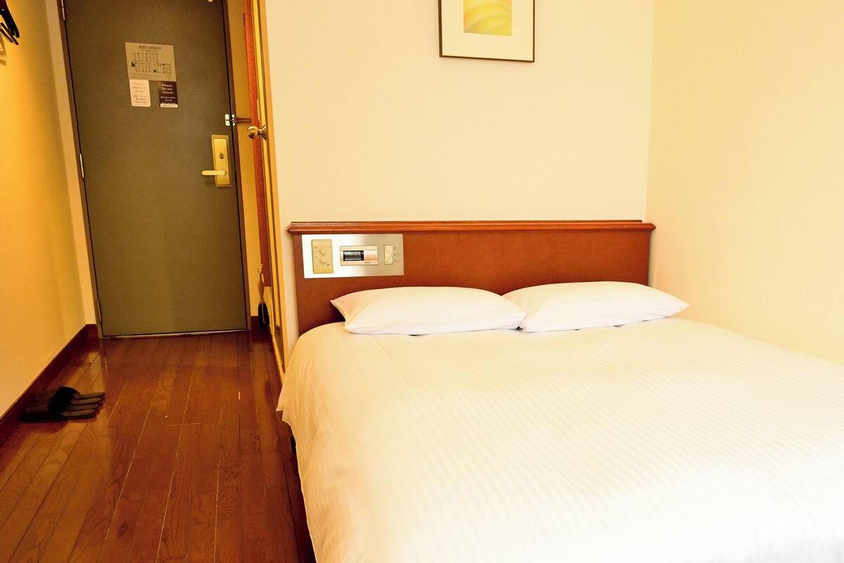Habitación de hotel con piso de madera, cama con ropa blanca, mesita de noche y termostato en la pared.