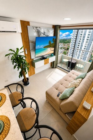 A modern living room with a flat-screen TV, a sofa, a dining area with chairs, a potted plant, and a balcony with a city view. - Caribbean Deluxe • Pool, Views & WiFi (Cartagena de Indias)