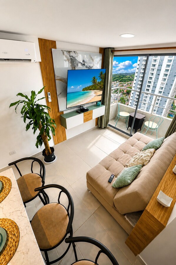 Ein modernes Wohnzimmer mit Fernseher, einer Couch, einem Essbereich mit Stühlen, einer Topfpflanze und einem Balkon mit Blick auf die Stadt.