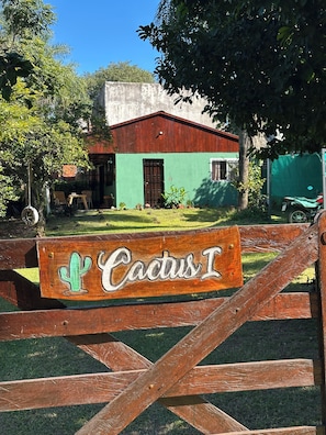 A wooden gate with a cactus sign, a green building with a red roof, and a concrete wall in the background.