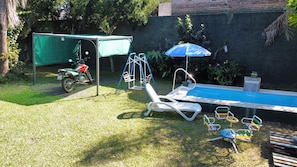 A backyard with a swimming pool, a covered area, a swing set, and a motorcycle.