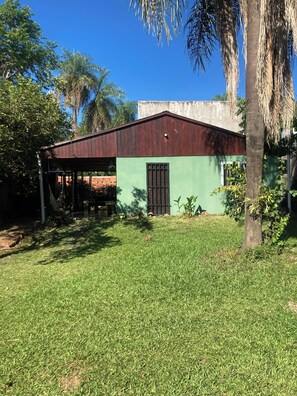 A small building with a green wall and a brown roof, surrounded by grass and palm trees.