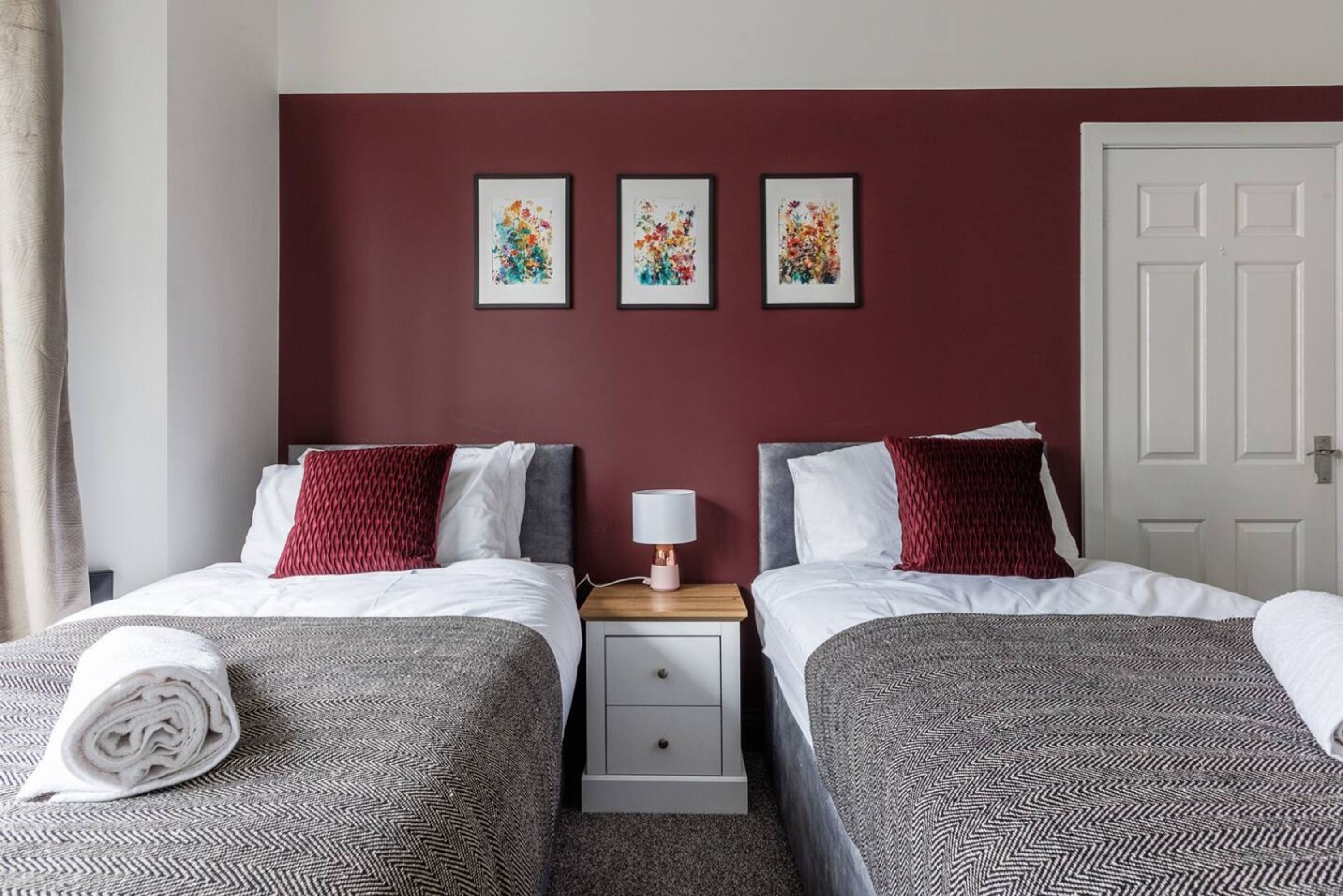 Une chambre d’hôtel avec deux lits, une table de chevet avec une lampe et trois œuvres d’art florales encadrées accrochées au mur.