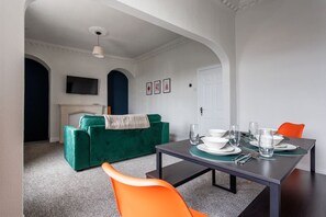 Ein Essbereich mit einem Tisch für vier Personen, einem grünen Sofa und einem an der Wand montierten Fernseher.