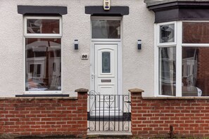 Ein zweigeschossiges Haus mit weißer Haustür, der Hausnummer '86' über der Tür und einem kleinen Balkon mit schwarzem schmiedeeisernen Geländer.