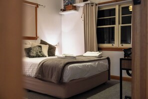 Un dormitori amb un llit, una taula de nit, una finestra amb cortines i una estant sobre el llit.