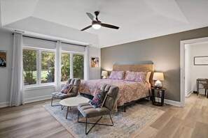 A bedroom with a bed, two armchairs, a ceiling fan, and large windows.