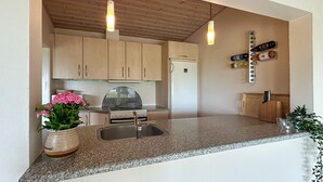 Een keuken met granieten aanrechtbladen, houten kastjes, een roestvrijstalen spoelbak, een koelkast en hanglampen.