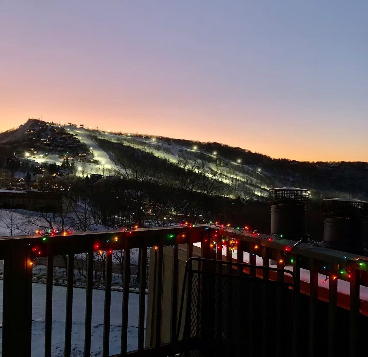A snowy landscape with a hill illuminated by streetlights, a metal railing, and a distant building.