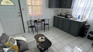 Een ruimtelijk ingerichte leefruimte met een bank, een klein tafeltje, een kitchenette met kastjes en een spoelbak, en een deur met een raam.
