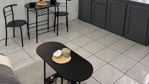 Een compacte keuken met grijze kastjes, een eettafel met stoelen, een kleine zwarte tafel met een kom en een plant, en een betegelde vloer.