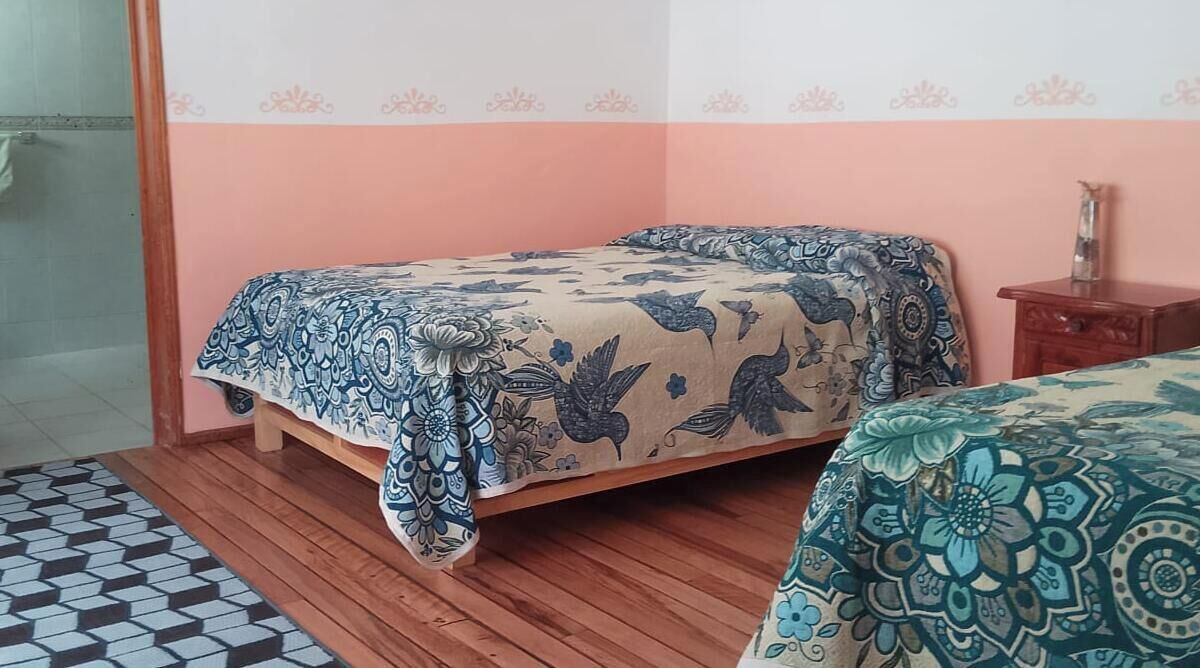 Un dormitorio con cama, suelo de madera, colcha estampada, mesita de noche y una silla.