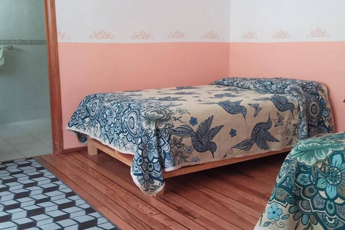 A bedroom with a bed, a wooden floor, a patterned bedspread, a nightstand, and a chair.
