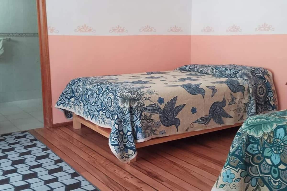 A bedroom with a bed, a wooden floor, a patterned bedspread, a nightstand, and a chair.