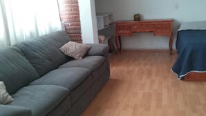 Ein Zimmer mit grauem Sofa, einem Holztisch, einem Bett mit blauer Bettdecke und einer Ziegelwand.