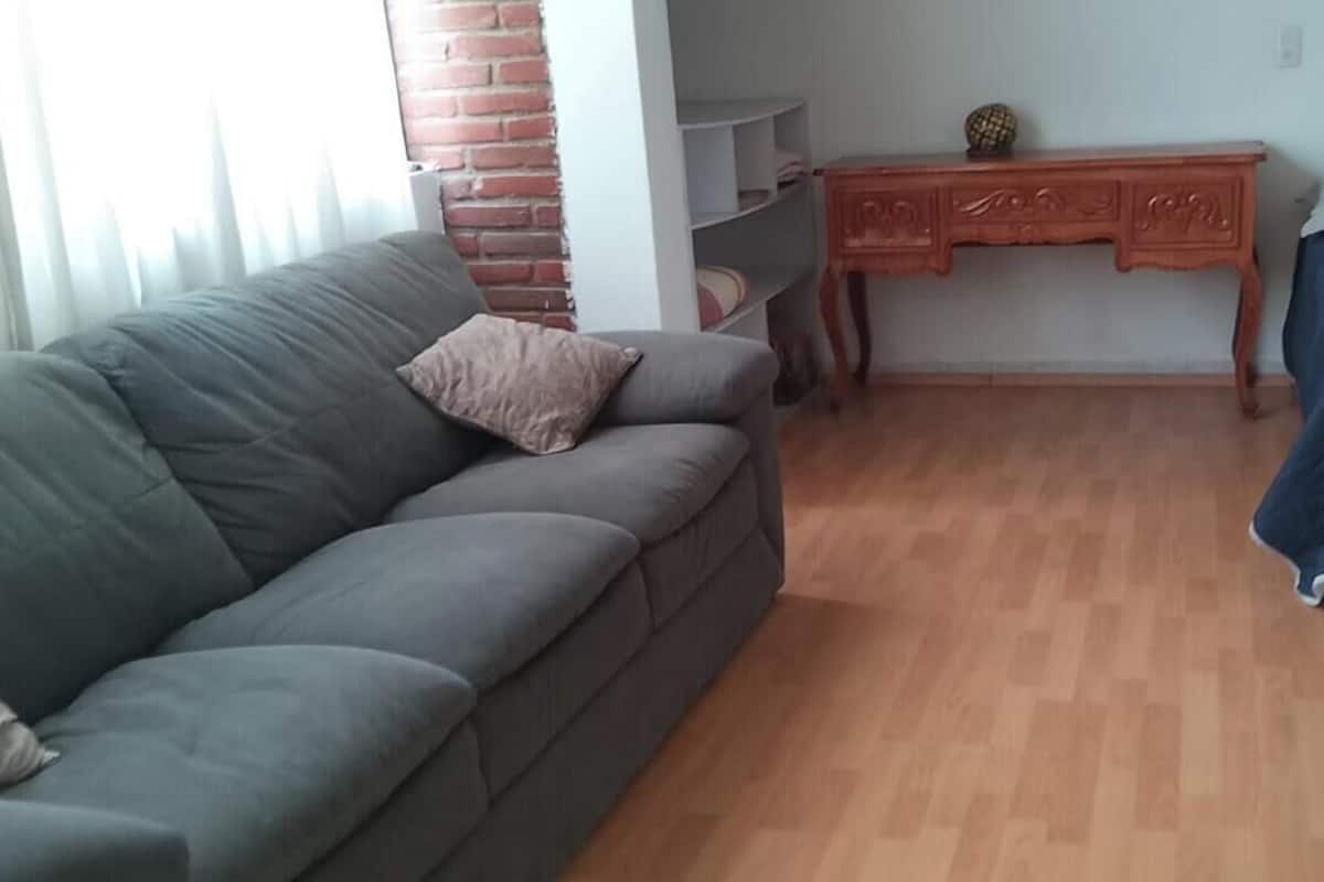A room with a grey sofa, a wooden desk, a bed with a blue cover, and a brick wall.