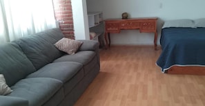 A room with a grey sofa, a wooden desk, a bed with a blue cover, and a brick wall.
