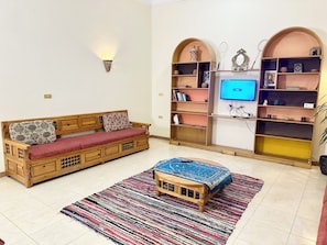 A living room with a wooden sofa, a television, shelves with decorative items, and a patterned rug.