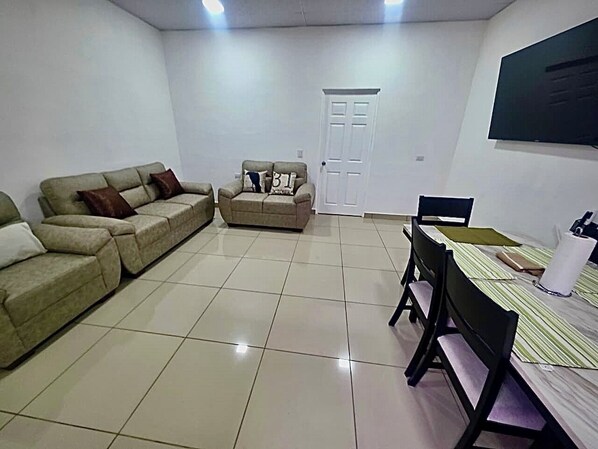 A living room with a sofa set, a dining table with chairs, a door, and a television mounted on the wall.