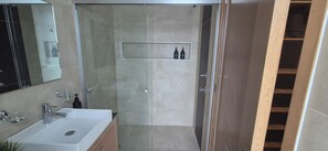 A modern bathroom with a glass-enclosed shower, a sink with a mirror, and a shelf with toiletries.