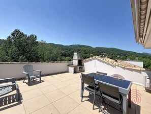En takterrass med ett bord och stolar, en grill och utsikt över grönskande kullar.